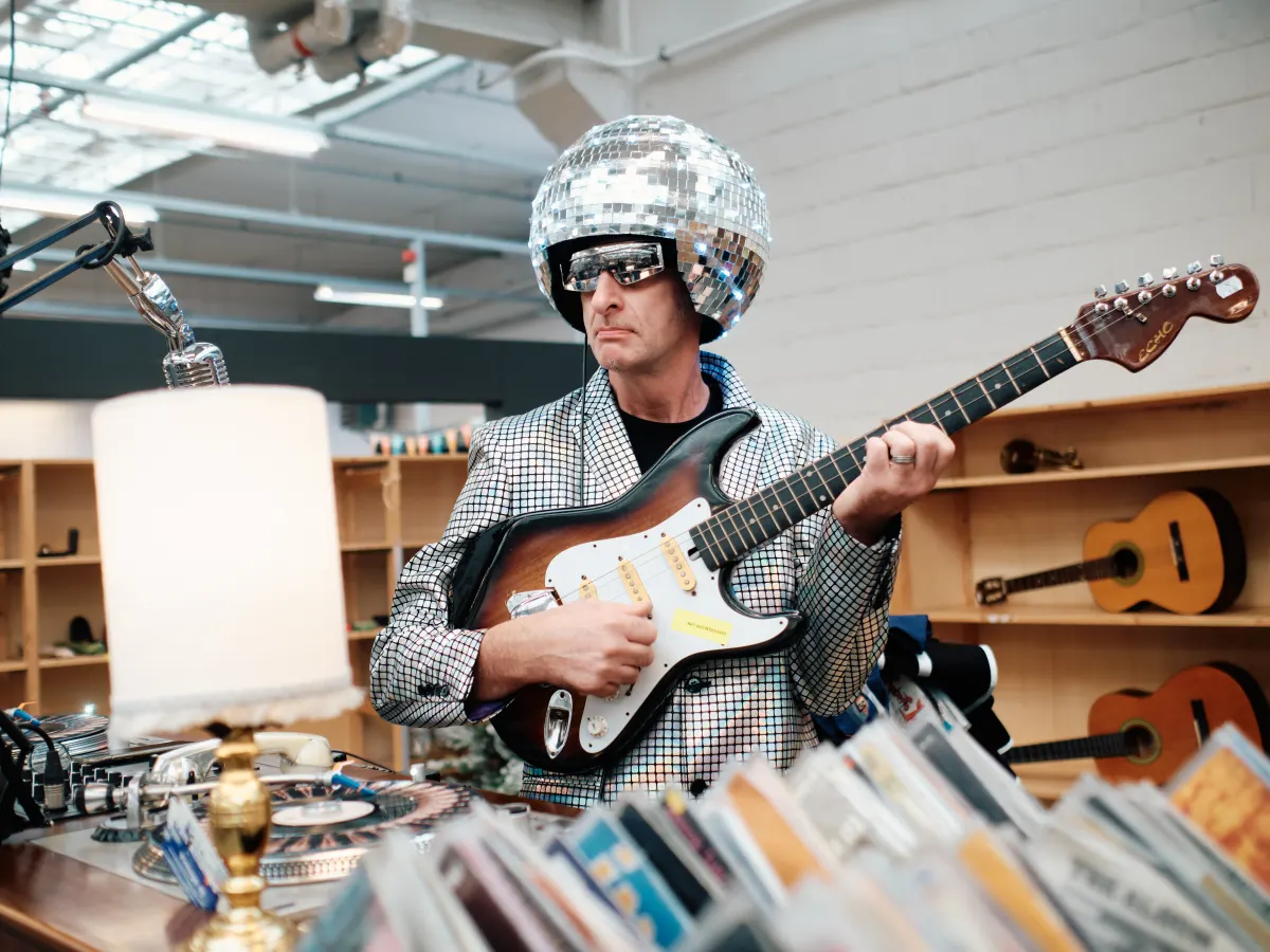 Een muzikant met discobolhelm speelt gitaar tussen de rekken van de Kringwinkel.